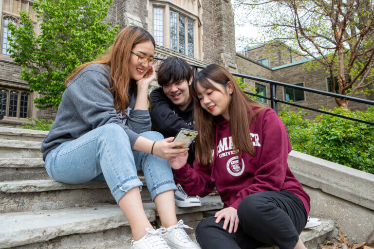 Three students sitting on stairs, looking at cell phone on McMaster campus.