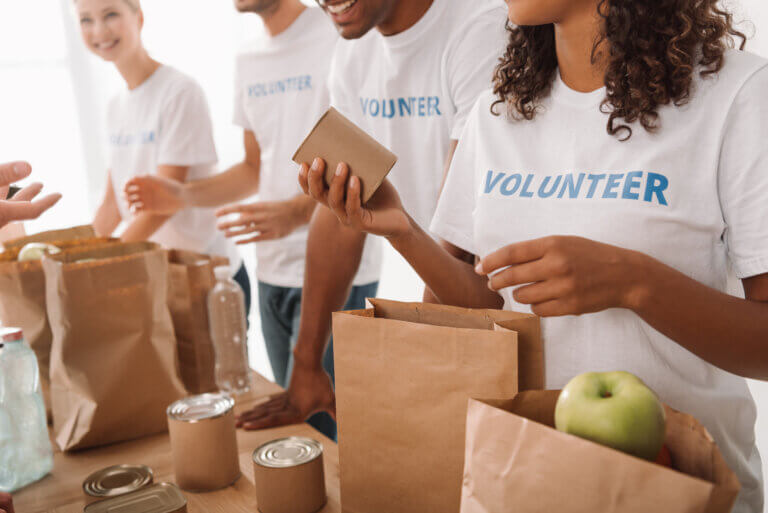Several volunteers wearing white T-shirts labeled “VOLUNTEER” pack food and supplies into brown paper bags at a table. The table contains canned goods, bottled water, and fresh produce, including apples, as volunteers assemble donation packages for a community food drive.