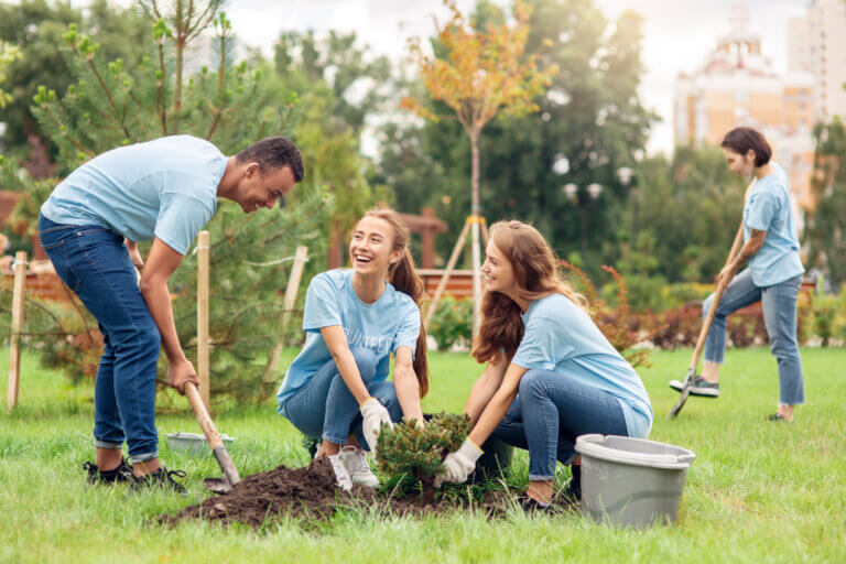 Four volunteers wearing light blue shirts work together in a park to plant a small shrub. One person digs a hole with a shovel while two others position the plant in the soil. Another volunteer uses a rake in the background. Green grass, young trees, and landscaped garden areas surround the group, with buildings visible in the distance on a bright day.