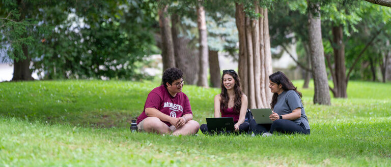Three students sit on green grass under trees, studying outdoors with laptops. They are smiling, creating a relaxed, collaborative atmosphere.