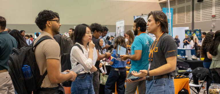 A crowded career fair or recruiting event inside a large convention hall. In the foreground, two attendees wearing backpacks and holding documents speak with a representative at an exhibitor booth. Additional attendees and recruiters are gathered around nearby tables and displays in the background, with banners, promotional materials, and lanyards visible throughout the busy event space.
