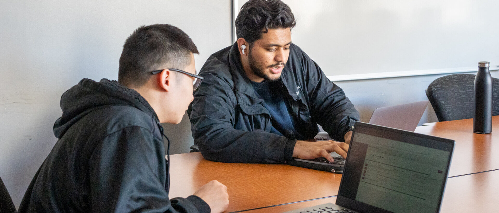 Two people in a meeting room focus on a laptop, discussing content visible on the screen. The mood is collaborative and engaged, with a modern, tech-savvy feel.