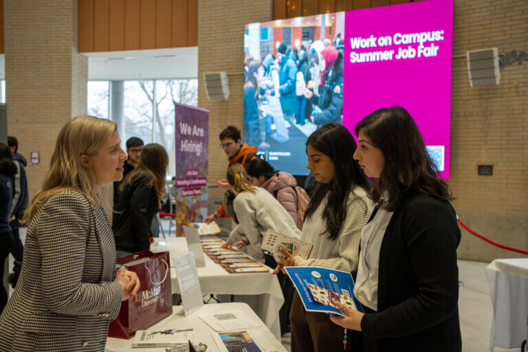 A woman at a job fair, interacting with two attendees holding pamphlets. A "We Are Hiring" sign and a large digital screen are visible in the background.