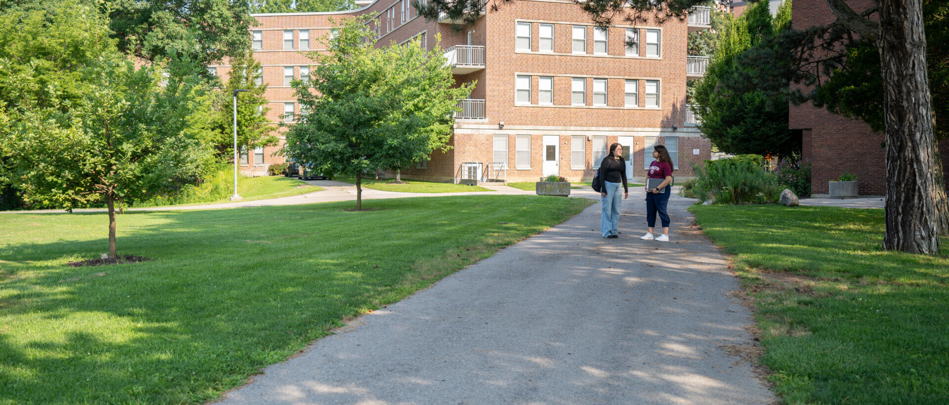 A path leads through a grassy area with trees toward a brick building. Two people walk along the path under a sunny, clear sky.