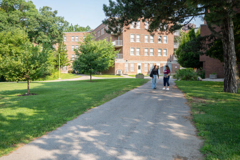 A path leads through a grassy area with trees toward a brick building. Two people walk along the path under a sunny, clear sky.