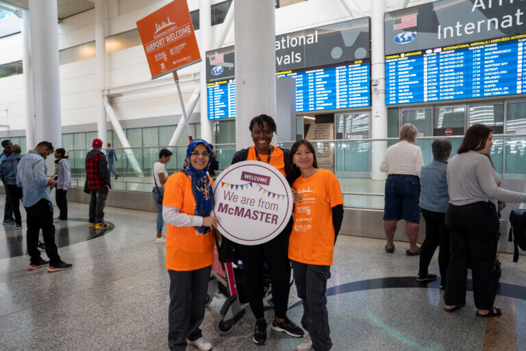 Three people in orange shirts hold a "We are from McMaster" sign at an airport