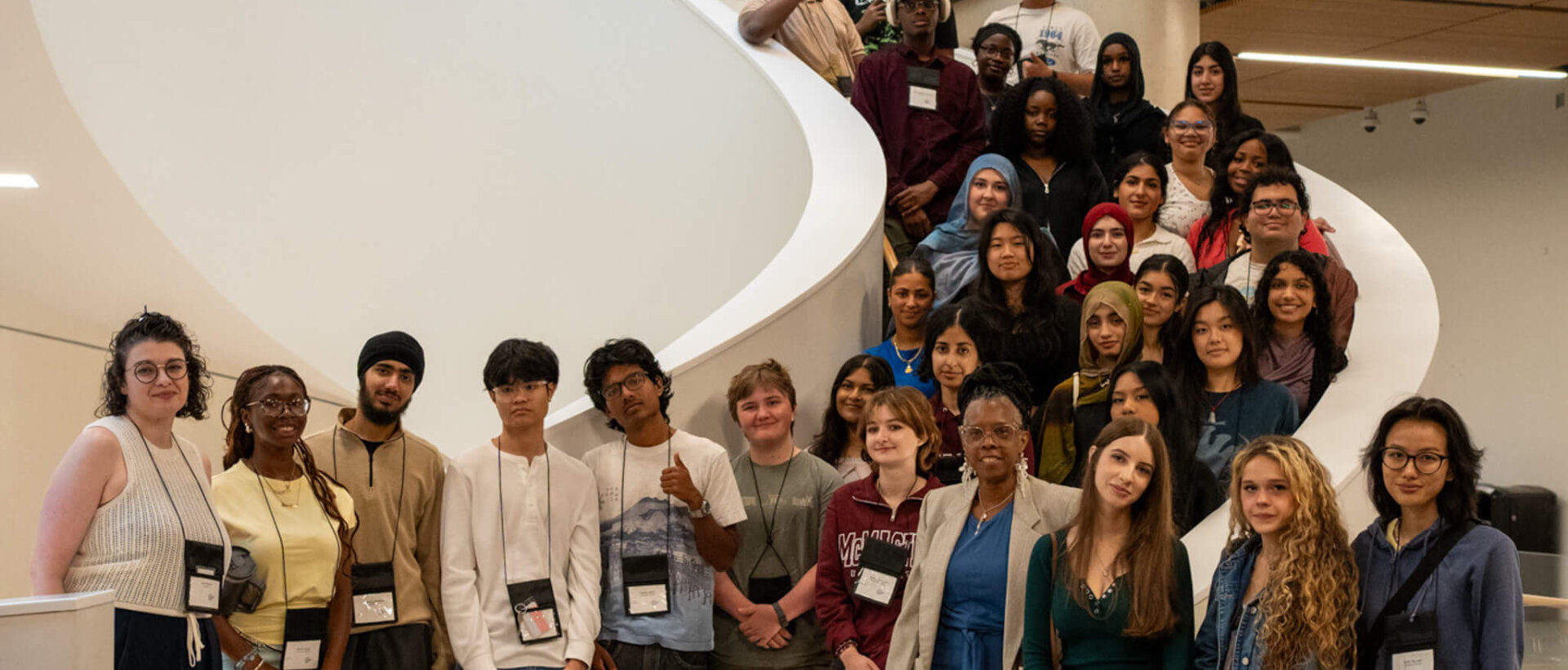 Large, diverse group of students and adults pose cheerfully on a sweeping indoor staircase in a bright, modern atrium.