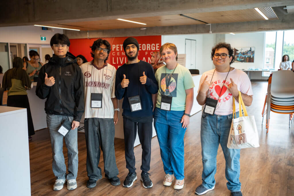 Five diverse young adults pose indoors at a lively event, smiling and giving thumbs-up while wearing badges and carrying a tote bag.