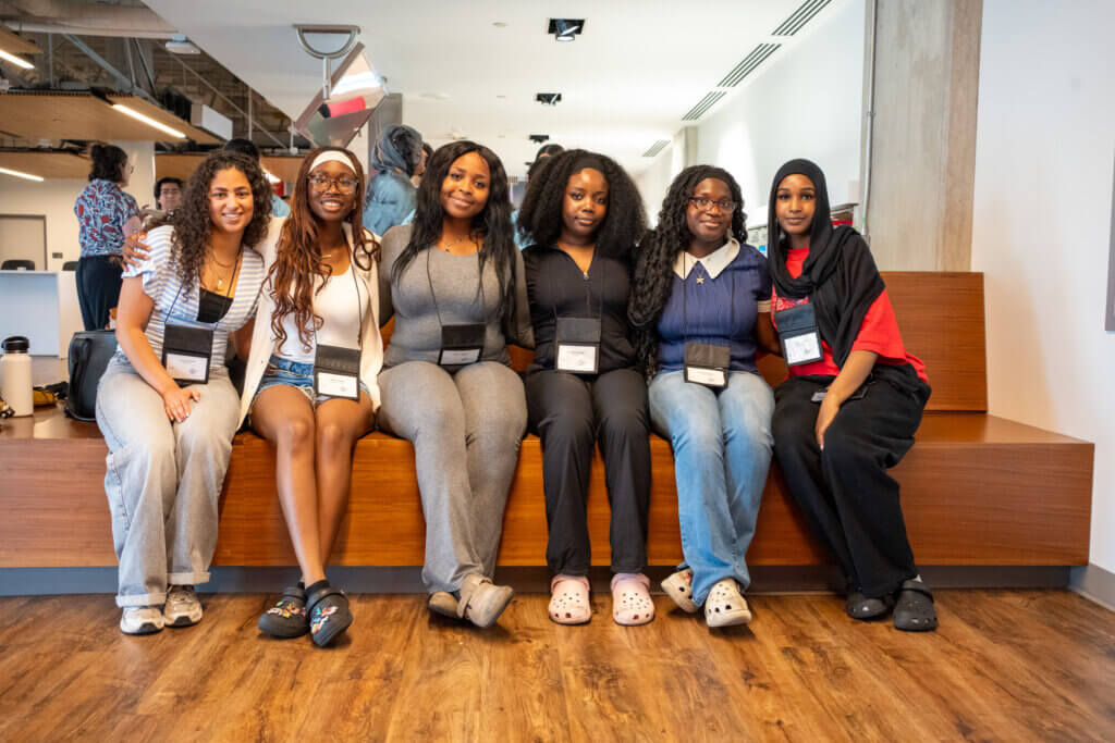 Six diverse women smile together while seated on a wooden bench in a bright office, conveying friendship, confidence, and camaraderie.
