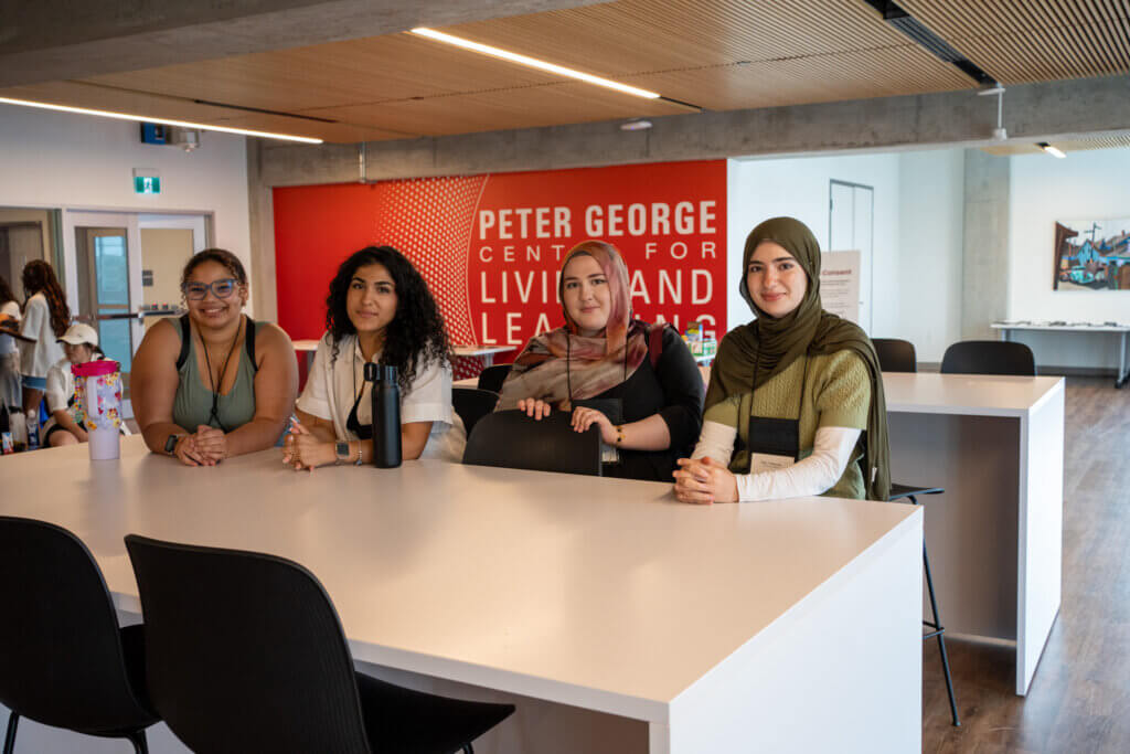 Four women sit around a table in a bright community center, smiling at the camera, with a Peter George Centre for Living and Learning sign behind them.