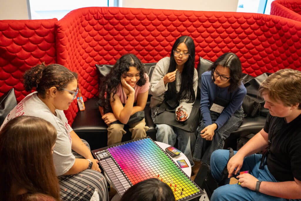 A diverse group of students gathers around a colorful board game, smiling and chatting in a cozy lounge with red cushioned walls.