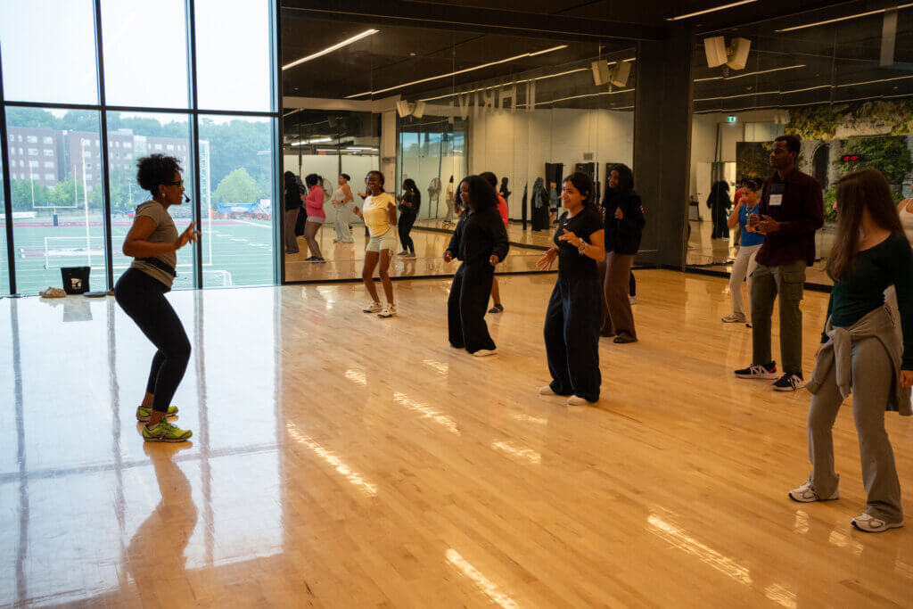 Dance instructor leads a diverse group in an energetic fitness class inside a mirrored studio with polished floors and city views.