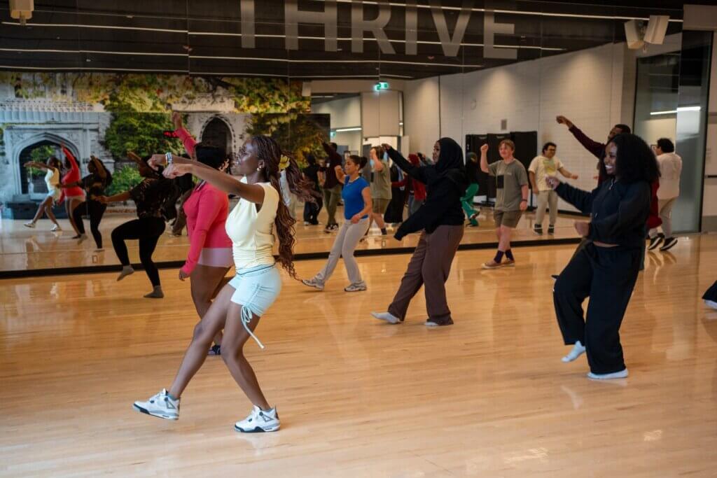 Diverse dancers follow a choreographed routine in a bright mirrored studio, moving energetically across the polished wood floor.