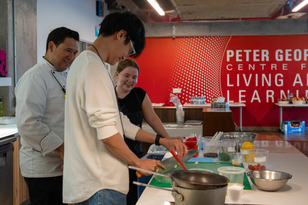 In a modern teaching kitchen, a young man stirs a pan while classmates watch and prepare food; a red culinary school sign fills the background.