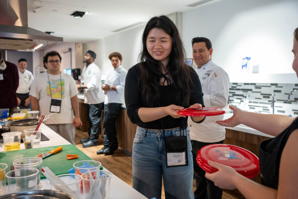 Smiling woman holds a red plate at a cooking event, surrounded by chefs and colorful ingredients in a bright kitchen; warm, social atmosphere.