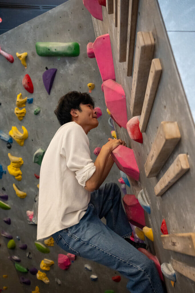 Focused climber in a white sweater and jeans grips pink holds on an indoor bouldering wall, surrounded by colorful routes and wooden volumes.