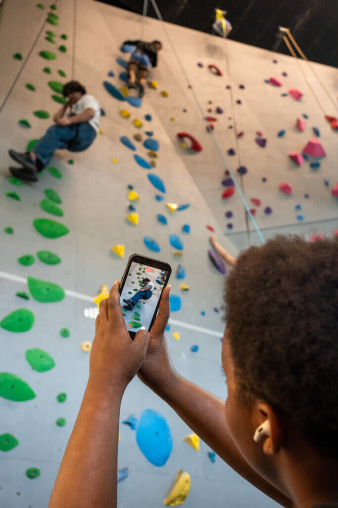 A person photographs climbers scaling a colorful indoor rock wall, capturing the energetic scene on a smartphone.
