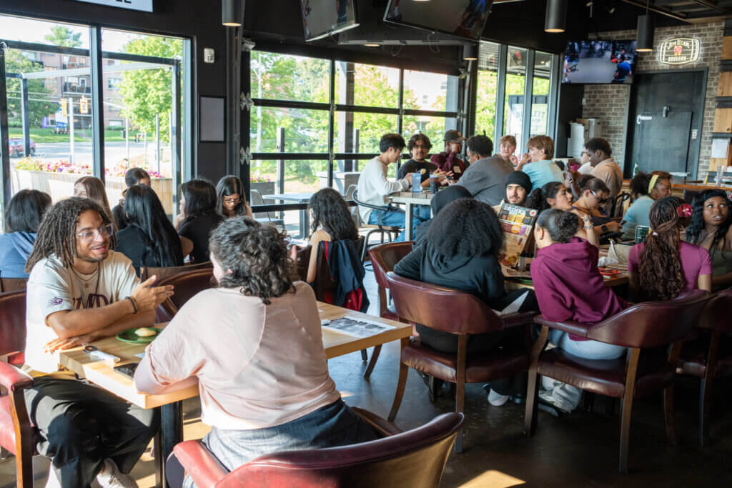 Lively café with diverse young adults dining and chatting at tables, sunlight streaming through large windows, creating a warm, social atmosphere.