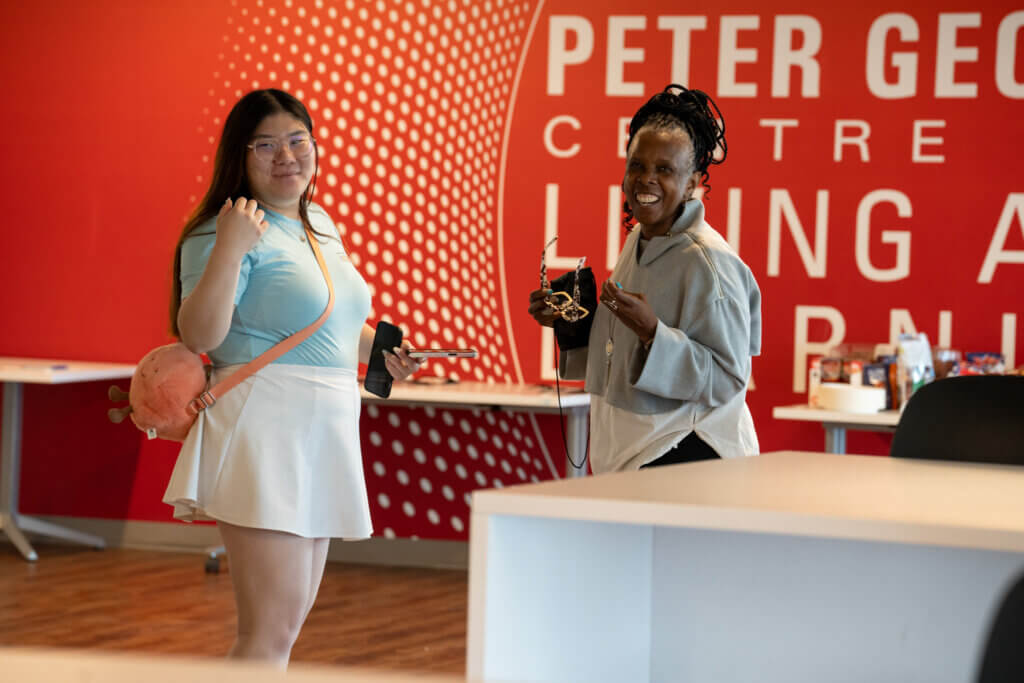 Two smiling women hold cameras in a bright event space, with a red wall displaying “Peter George Centre” behind them.