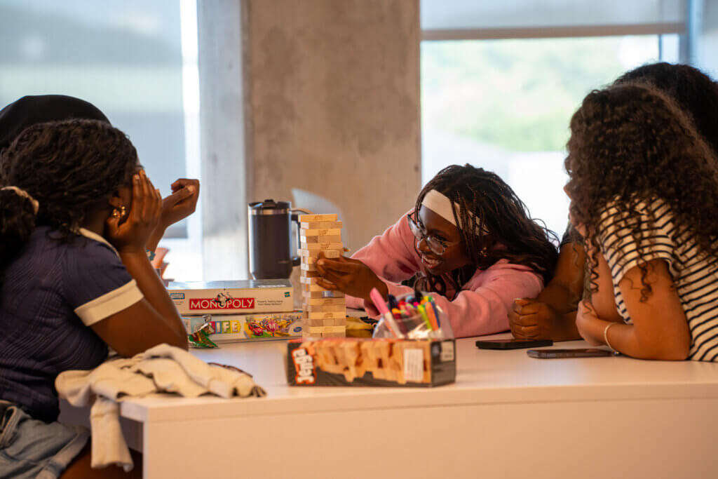 Three friends play Jenga at a table, laughing and focused as one carefully removes a block; Monopoly and colorful pens sit nearby.