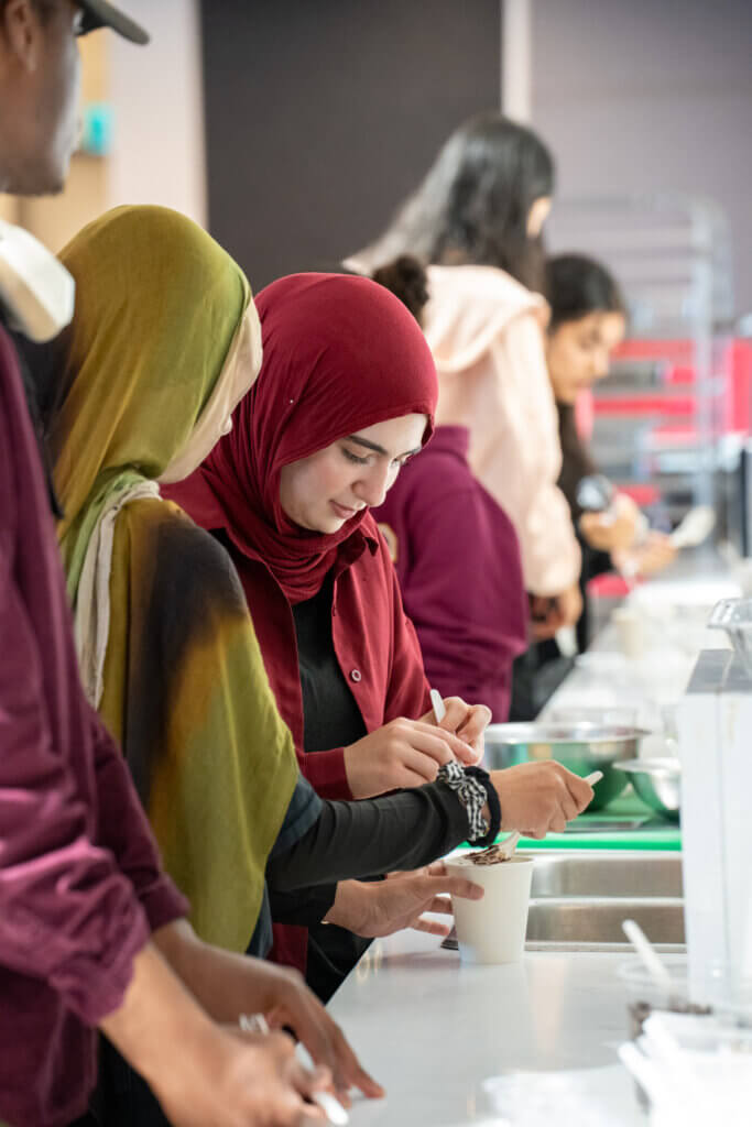People in hijabs and casual clothing prepare food together at a kitchen counter, creating a warm, collaborative community atmosphere.