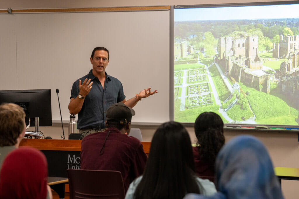 Lecturer gestures during a classroom presentation as students watch a projected image of historic castle ruins and gardens, creating an engaging academic atmosphere.
