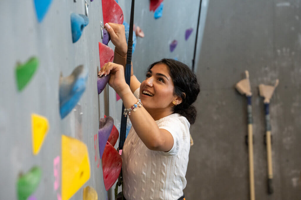 Smiling woman climbs an indoor rock wall, gripping colorful holds, while climbing gear and walking poles hang on the gray wall behind her.