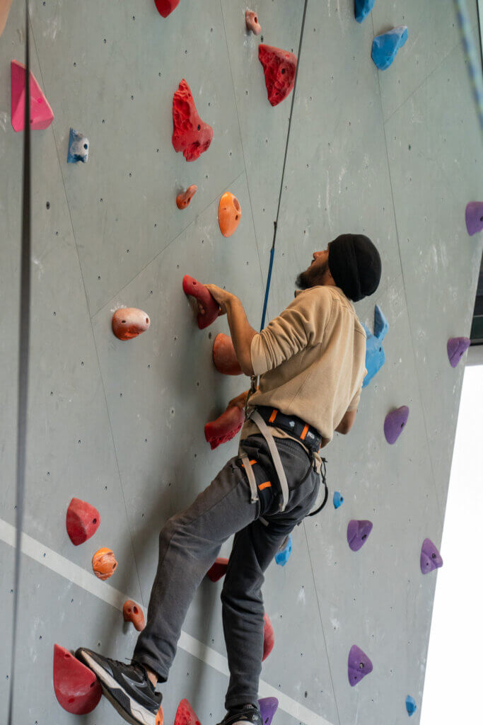 Indoor rock climber in a harness grips colorful holds on a tall gray climbing wall, reaching upward with focused determination.