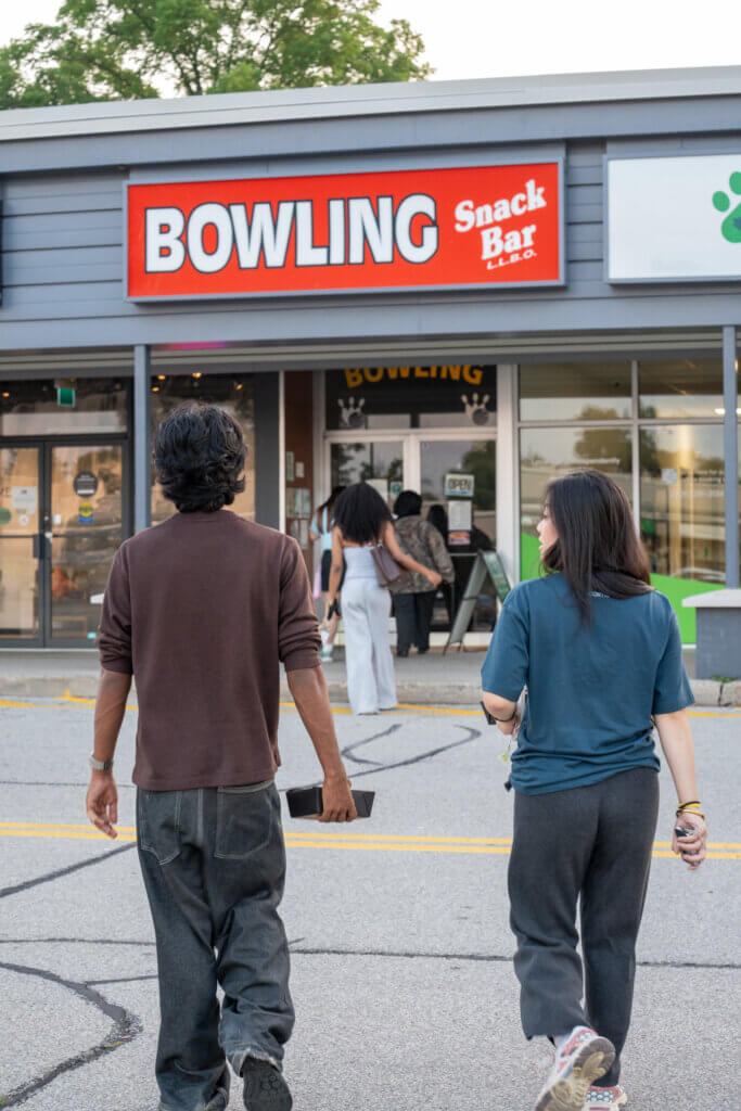 Two adults walk toward a bowling alley entrance beneath a bright red “BOWLING Snack Bar” sign, creating a casual, inviting atmosphere.