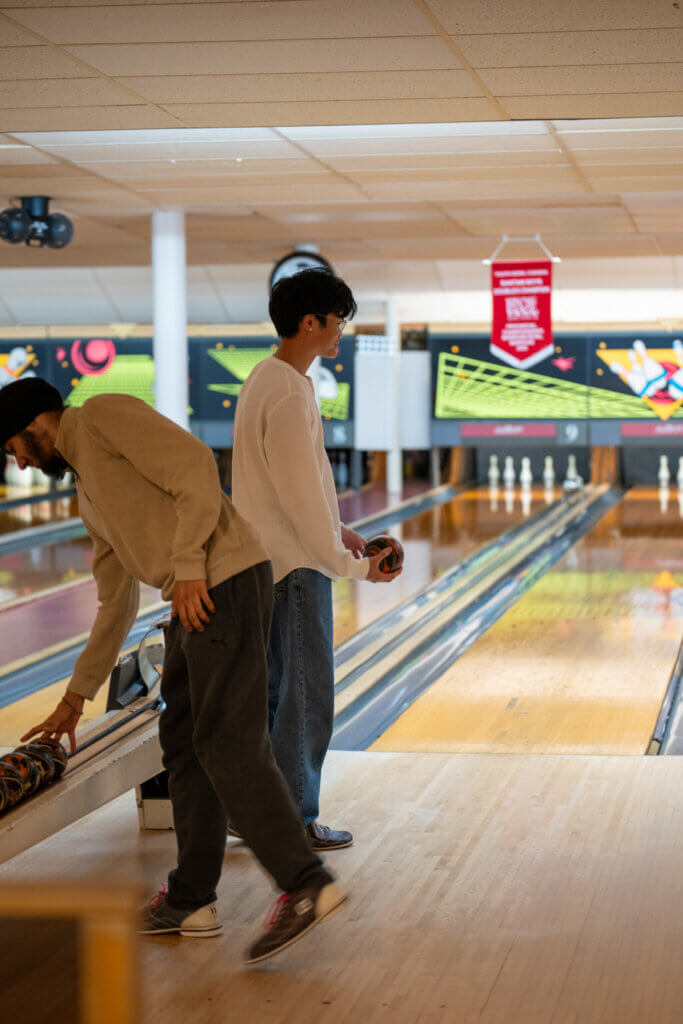 Two bowlers at a bowling alley, one bending to pick up a ball while the other prepares to roll, creating a relaxed, focused atmosphere.