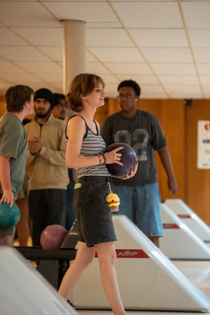 Young woman carrying a bowling ball walks past lanes as friends watch in a bright bowling alley, creating a relaxed, social atmosphere.