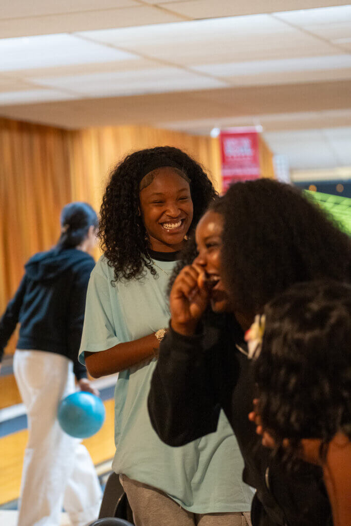 Two Black women laugh and whisper at a bowling alley; one wears a mint shirt, while a bowler and blue ball appear behind them.