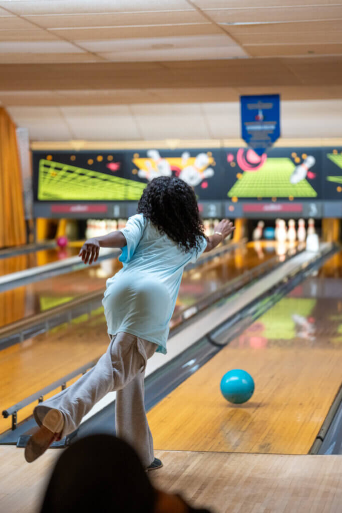Bowler in a light blue shirt releases a blue ball down a brightly lit bowling lane, balancing on one leg as pins await.