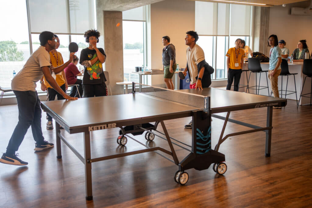 Two people playing ping pong in a modern room with a wooden floor. A group of onlookers, casually dressed, watch and chat in the background.
