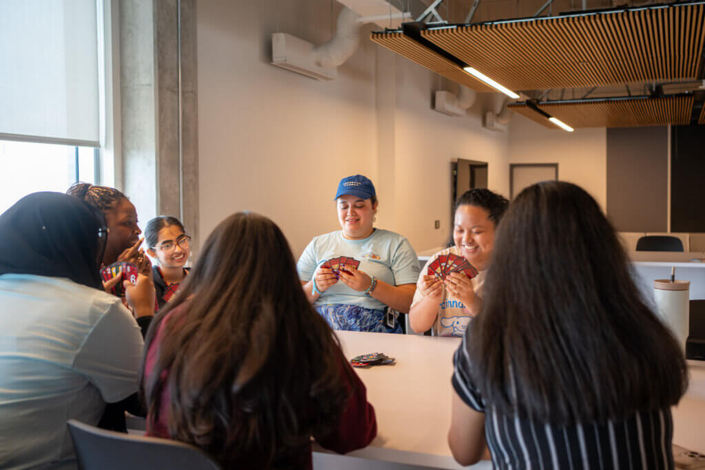 A group of seven people sit around a table playing a card game, smiling and engaged. The room is bright with natural light and has a modern ceiling design.