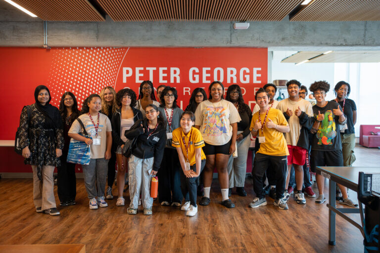 A diverse group of students stands smiling in front of a wall with "Peter George" text, conveying a sense of unity and positivity in a modern room.