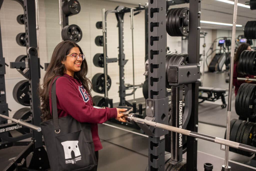 A young woman smiles while adjusting weights on a squat rack in a gym. She wears a maroon sweatshirt and a black tote bag, creating a relaxed and positive atmosphere.