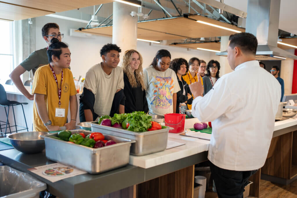 A group of attentive students faces a chef in a modern kitchen. Fresh vegetables are on the counter, creating an educational and engaging atmosphere.