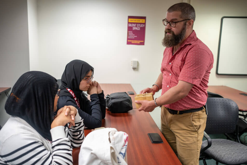 Man in a red shirt shows a small wooden object to two seated women in hijabs at a table labeled "Silent Study Space." They appear attentive.
