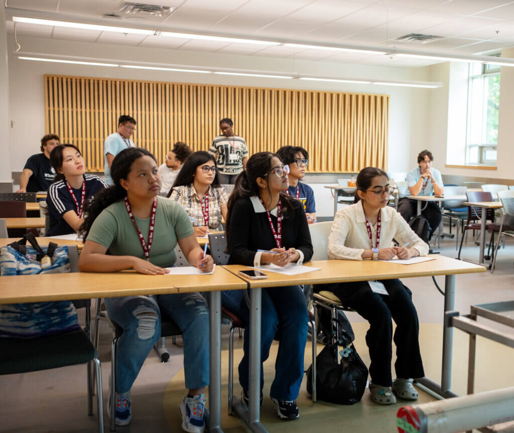 A diverse group of students attentively listen in a classroom. They sit at desks with notebooks, wearing lanyards, conveying focus and engagement.