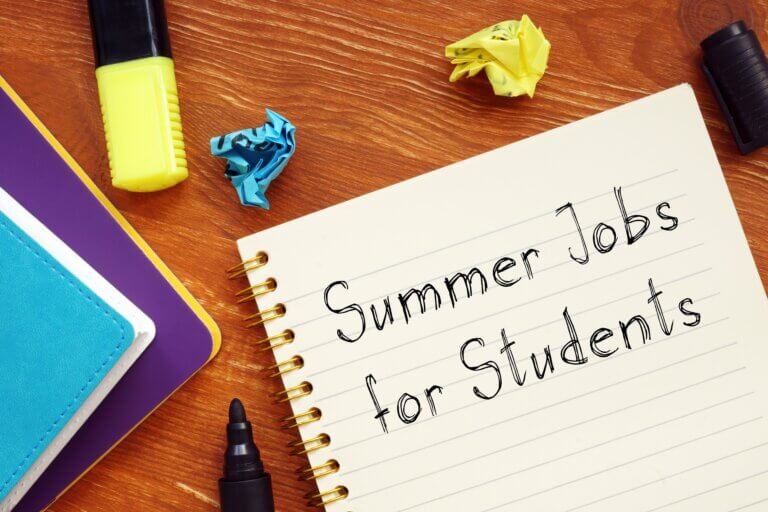 Spiral notebook reading “Summer Jobs for Students” on a wooden desk, surrounded by colorful notebooks, markers, and crumpled paper.
