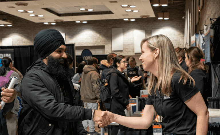 At a busy convention, a Sikh man in a black turban and a blonde woman smile and shake hands, while attendees mingle among booths behind them.
