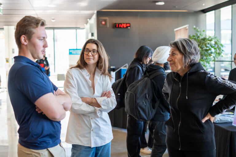 Three people stand in an indoor setting, engaged in conversation, arms crossed. They look attentive and thoughtful. A clock reads 11:26, and others are in the background.