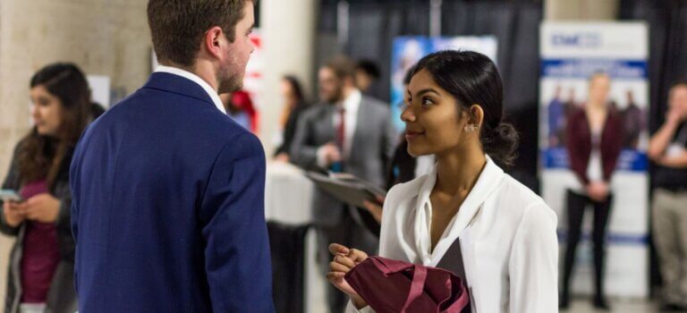 A woman in a white blouse holds a folder, engaged in conversation with a man in a blue suit. Blurry people and professional banners are in the background, suggesting a networking event.