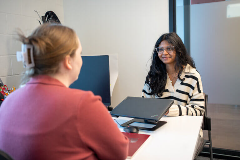 Two women are sitting across a desk in an office setting, engaged in conversation. One faces forward with a smile, while the other listens attentively.