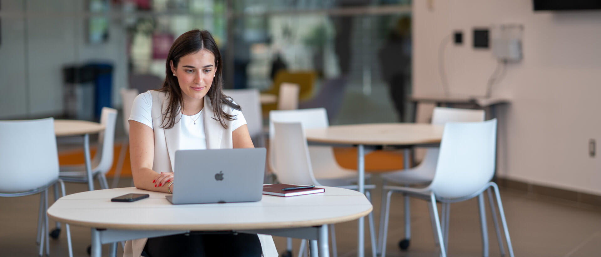 A woman sits at a round table in a modern, bright office space, focused on her laptop. She is surrounded by empty chairs, exuding a professional ambiance.