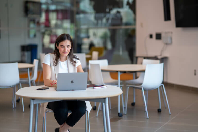 A woman sits at a round table in a modern, bright office space, focused on her laptop. She is surrounded by empty chairs, exuding a professional ambiance.