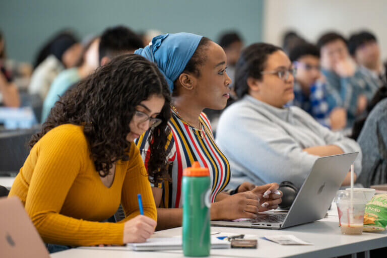 Students attentively listen in a classroom, with focus on a person taking notes and another using a laptop. The atmosphere is focused and studious.