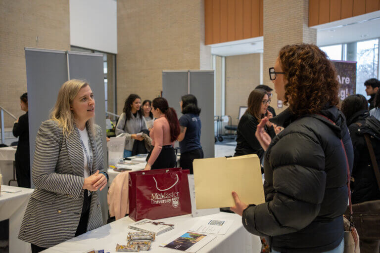 A woman in a checkered blazer talks to a person holding documents at a McMaster University booth in a bustling indoor event. The atmosphere is engaging.