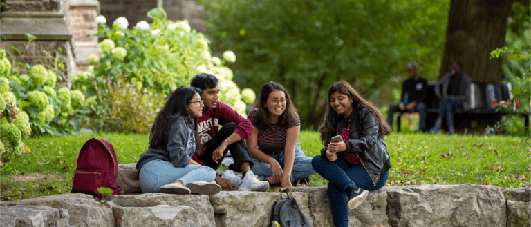 Four students sit on a stone ledge in a park, laughing and chatting. They are surrounded by greenery, enjoying a relaxed, cheerful atmosphere.